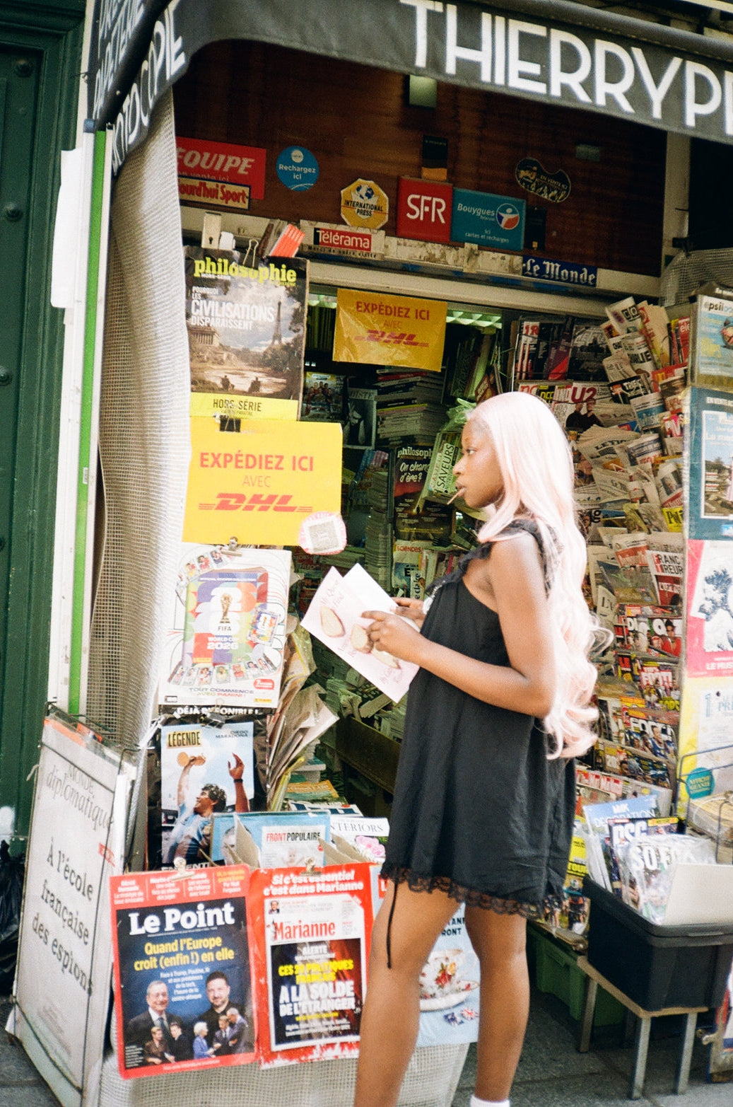 A woman with long light pink hair wearing a black dress stands in a crowded newspaper stand, looking down at a magazine or booklet she is holding.